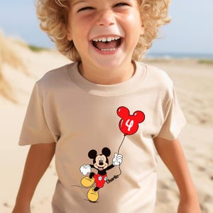 May include: A beige t-shirt with a Mickey Mouse cartoon holding a red balloon with the number 4. The shirt has a personalized name below the character. The child is smiling on a sandy beach.