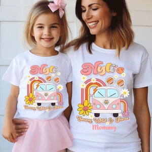 May include: Matching white t-shirts featuring a pink and white retro van, rainbow, and flowers. The child's shirt reads "Nyla Young Wild & Three". The adult shirt reads "Nyla's Young Wild & Three Mommy".