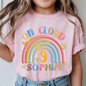 May include: Pink t-shirt with a colorful rainbow design and the text "ON CLOUD 9 SOPHIA". The number 9 is in the center of the rainbow. Gold heart accents are on the shirt.