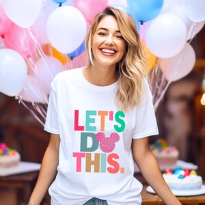 May include: White t-shirt with the text "LET'S DO THIS" in colorful block letters and a pink glitter Mickey Mouse head. The shirt is worn by a person smiling in front of balloons and cakes.