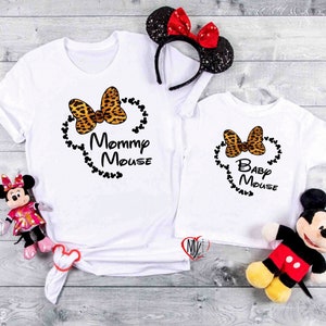 May include: Two white t-shirts with matching designs. Each shirt features a leopard print bow, a string of black Mickey Mouse heads, and the words "Mommy Mouse" and "Baby Mouse". A Minnie Mouse headband and plush toys are also visible.