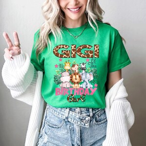 May include: A bright green t-shirt featuring a graphic print with the text GIGI in a brown giraffe print pattern. Below, a collection of cartoon safari animals including a giraffe, monkey, zebra, hippo, lion, and elephant with pink stars.
