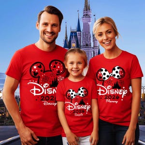 May include: Three red short-sleeved t-shirts featuring a black Mickey or Minnie Mouse ear silhouette graphic with white polka dots, fireworks, a castle outline, and the text Disney 2026. The shirts are personalised with Daddy, Mommy, and Sophia.