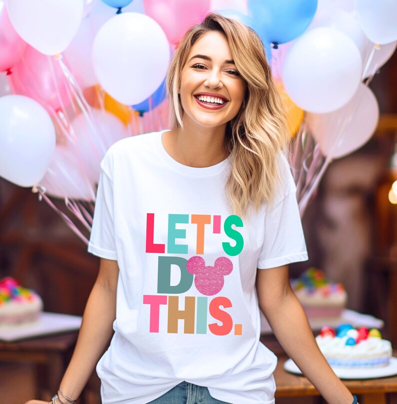 May include: White t-shirt with the text "LET'S DO THIS" in colorful block letters and a pink glitter Mickey Mouse head. The shirt is worn by a person smiling in front of balloons and cakes.