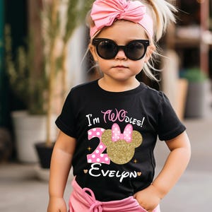 Puede incluir: Una camiseta negra con un n&uacute;mero "2" de purpurina dorada y una cabeza de Minnie Mouse rosa con un lazo rosa. La camiseta dice "I'm Twodles!" y "Evelyn" en letras doradas de purpurina.