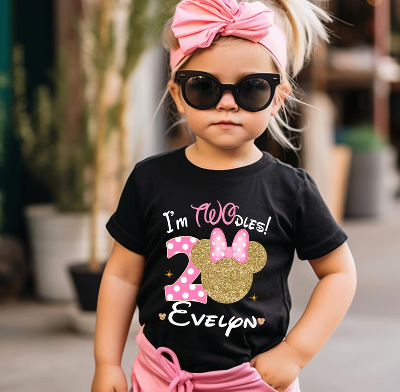 Puede incluir: Una camiseta negra con un n&uacute;mero "2" de purpurina dorada y una cabeza de Minnie Mouse rosa con un lazo rosa. La camiseta dice "I'm Twodles!" y "Evelyn" en letras doradas de purpurina.