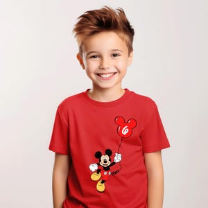May include: A red t-shirt with a Mickey Mouse cartoon design holding a red balloon with the number 6. The short-sleeved shirt is worn by a smiling child.