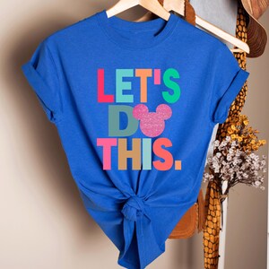 May include: A blue t-shirt with the phrase "LET'S DO THIS" in colorful block letters. The word "DO" features a pink glittery Mickey Mouse head. The shirt is knotted at the waist and hangs on a wooden hanger.