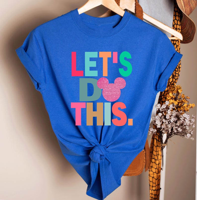 May include: A blue t-shirt with the phrase "LET'S DO THIS" in colorful block letters. The word "DO" features a pink glittery Mickey Mouse head. The shirt is knotted at the waist and hangs on a wooden hanger.