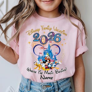 May include: Pink t-shirt featuring a Disney Family Vacation design for 2026. The design includes the iconic Mickey Mouse ears, a castle, fireworks, and Disney characters. The text "Where the Magic Begins!" and a space for a name are also included.
