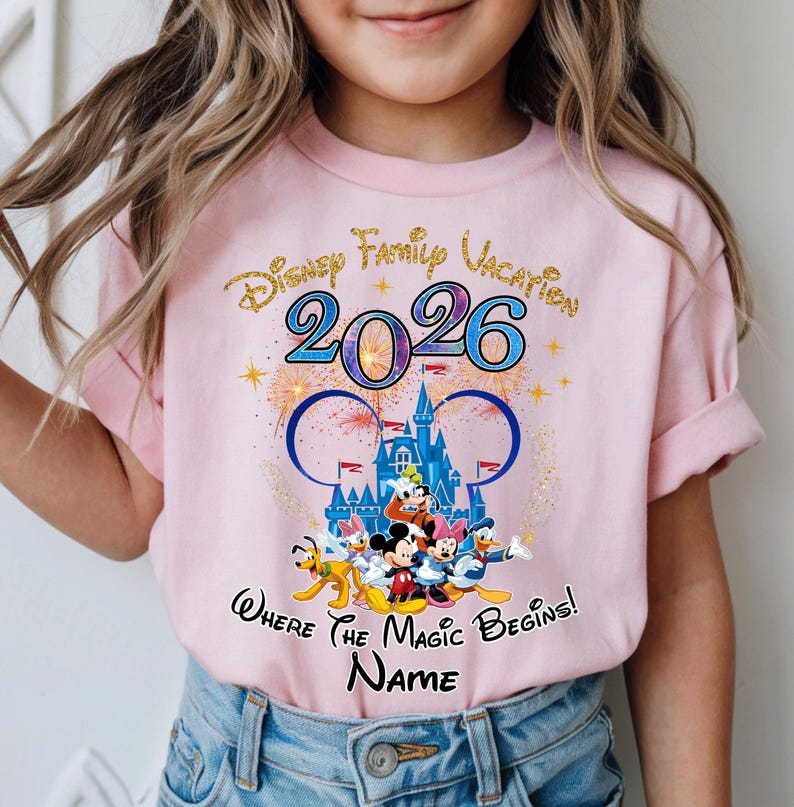 May include: Pink t-shirt featuring a Disney Family Vacation design for 2026. The design includes the iconic Mickey Mouse ears, a castle, fireworks, and Disney characters. The text "Where the Magic Begins!" and a space for a name are also included.