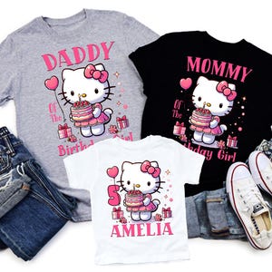 May include: Three t-shirts featuring a Hello Kitty design, ideal for a birthday. The shirts are grey, black, and white, with pink text and graphics. The grey shirt says "Daddy of the Birthday Girl." The black shirt says "Mommy of the Birthday Girl." The white shirt says "5 Amelia."