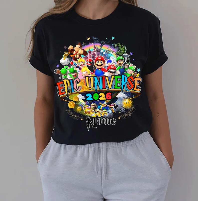 Pu&ograve; includere: T-shirt nera con una grafica colorata. Il design include il testo "Epic Universe 2026" e vari personaggi dei cartoni animati. Sono presenti anche un arcobaleno, stelle e monete d'oro. &Egrave; inclusa la parola "Name".