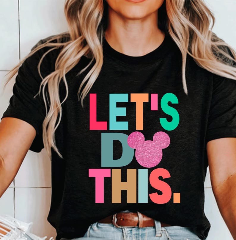 May include: A black t-shirt with the words 'LET'S DO THIS' in colorful letters. The word 'DO' has a pink glitter Mickey Mouse head over it.