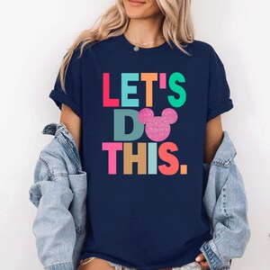 May include: Navy blue t-shirt with the text "LET'S DO THIS" in colorful block letters and a glittery pink Mickey Mouse head. The shirt is a casual style, suitable for everyday wear.