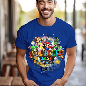 Pu&ograve; includere: T-shirt blu reale con una grafica colorata di personaggi di videogiochi. La grafica include il testo "EPIC UNIVERSE 2026" e uno spazio per un nome. Il design &egrave; circondato da stelle e monete d'oro.