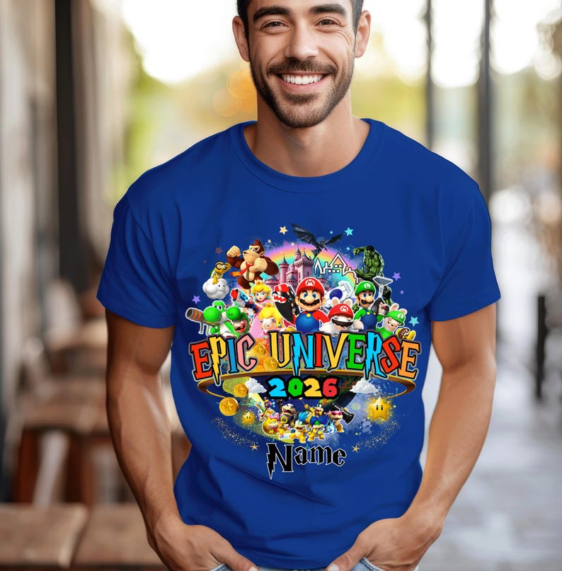 Pu&ograve; includere: T-shirt blu reale con una grafica colorata di personaggi di videogiochi. La grafica include il testo "EPIC UNIVERSE 2026" e uno spazio per un nome. Il design &egrave; circondato da stelle e monete d'oro.