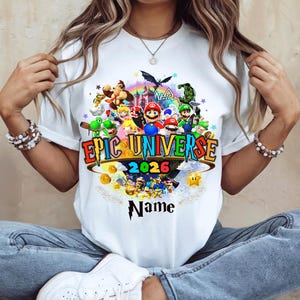 Pu&ograve; includere: T-shirt bianca con una grafica colorata. Il design include il testo "Epic Universe 2026" e vari personaggi dei cartoni animati. Sono presenti anche un arcobaleno, un castello e monete d'oro. La parola "Name" &egrave; sotto la grafica.