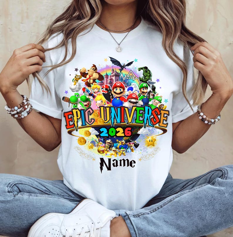 Pu&ograve; includere: T-shirt bianca con una grafica colorata. Il design include il testo "Epic Universe 2026" e vari personaggi dei cartoni animati. Sono presenti anche un arcobaleno, un castello e monete d'oro. La parola "Name" &egrave; sotto la grafica.