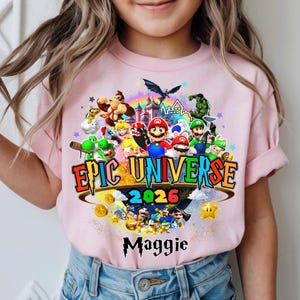 Pu&ograve; includere: T-shirt rosa chiaro con una grafica colorata di personaggi di videogiochi, la scritta "EPIC UNIVERSE 2026" e il nome "Maggie". Il design include un arcobaleno, stelle e monete d'oro.