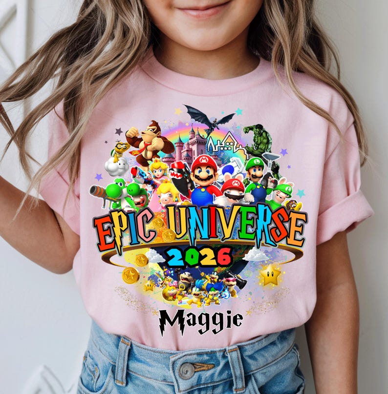 Pu&ograve; includere: T-shirt rosa chiaro con una grafica colorata di personaggi di videogiochi, la scritta "EPIC UNIVERSE 2026" e il nome "Maggie". Il design include un arcobaleno, stelle e monete d'oro.