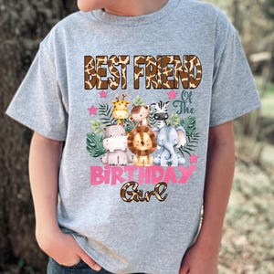 May include: A heather gray t-shirt featuring a graphic of a giraffe, monkey, zebra, hippo, lion, and elephant surrounded by green leaves and pink stars. The text reads BEST FRIEND Of The BIRTHDAY Girl in brown and pink lettering.