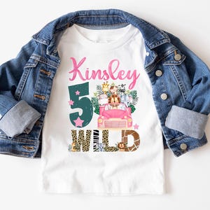 May include: A white t-shirt featuring the name Kinsley in pink script, a large dark green number 5, and a pink car filled with illustrated safari animals. The word WILD is printed below in animal print patterns, layered under a blue denim jacket.