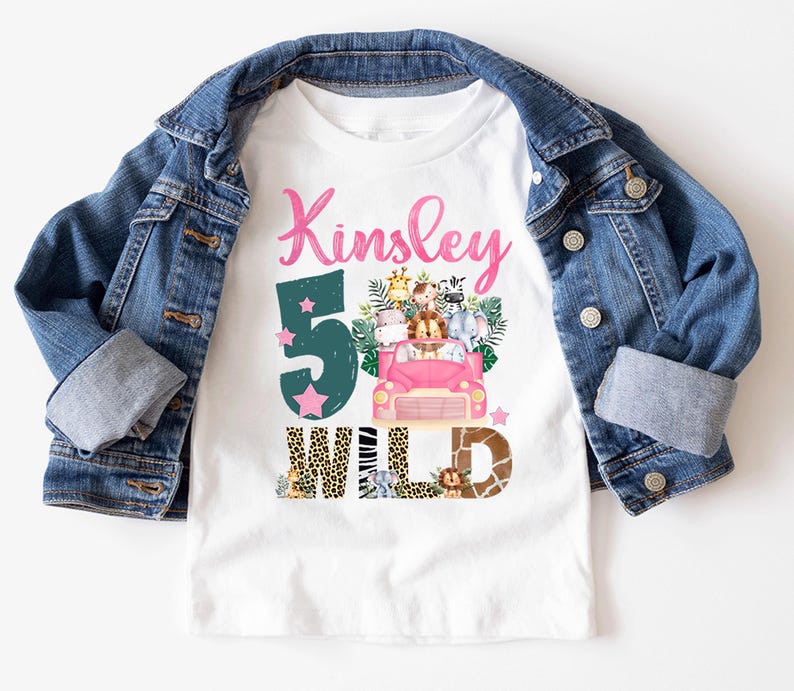 May include: A white t-shirt featuring the name Kinsley in pink script, a large dark green number 5, and a pink car filled with illustrated safari animals. The word WILD is printed below in animal print patterns, layered under a blue denim jacket.