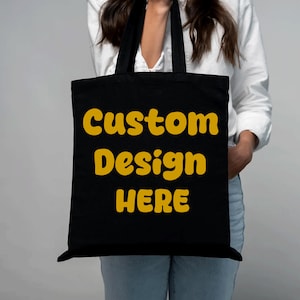 Puede incluir: Bolso tote negro con el texto "Custom Design HERE" en amarillo. El bolso tiene asas negras y lo sostiene una persona que lleva una camisa blanca y vaqueros azules. El bolso está hecho de un material resistente y está diseñado para transportar objetos.