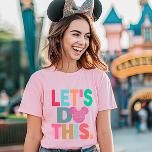May include: A pink t-shirt with the text "LET'S DO THIS" in colorful block letters and a glittery mouse head. The person is wearing black mouse ears with a patterned bow.