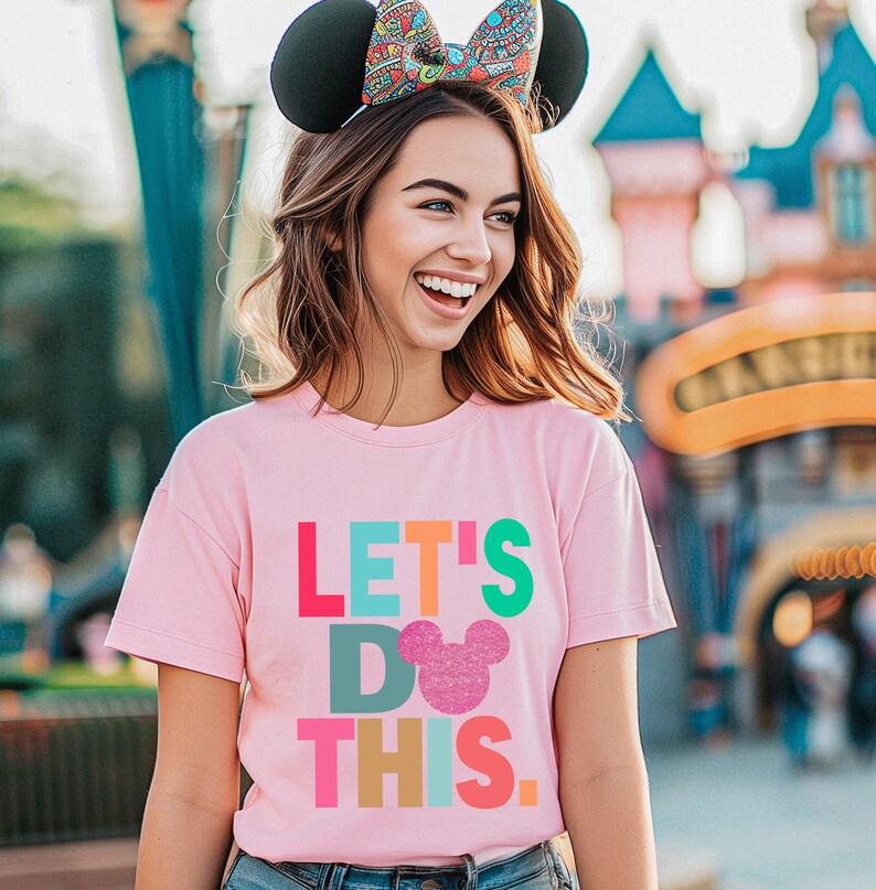 May include: A pink t-shirt with the text "LET'S DO THIS" in colorful block letters and a glittery mouse head. The person is wearing black mouse ears with a patterned bow.
