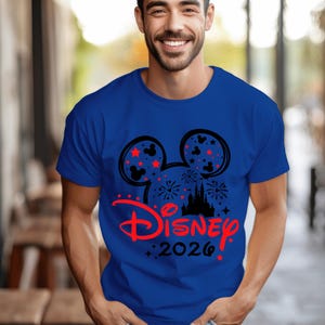 May include: A royal blue short-sleeved t-shirt featuring a black Mickey Mouse ear silhouette graphic filled with fireworks and small red stars. Below the ears, the word Disney is printed in red script, followed by the black text 2026.