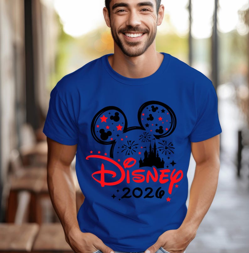 May include: A royal blue short-sleeved t-shirt featuring a black Mickey Mouse ear silhouette graphic filled with fireworks and small red stars. Below the ears, the word Disney is printed in red script, followed by the black text 2026.