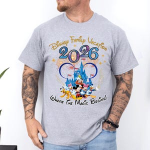 May include: A light gray t-shirt featuring a colorful graphic design. The design includes the text "Disney Family Vacation 2026" and a depiction of Disney characters in front of a castle. The phrase "Where the Magic Begins!" is also included.