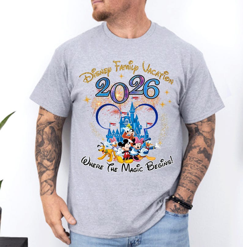May include: A light gray t-shirt featuring a colorful graphic design. The design includes the text "Disney Family Vacation 2026" and a depiction of Disney characters in front of a castle. The phrase "Where the Magic Begins!" is also included.
