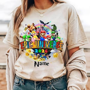 Pu&ograve; includere: T-shirt beige con una grafica colorata di personaggi di videogiochi, un arcobaleno e la scritta "Epic Universe 2026". Il design include uno spazio per un nome personalizzato. La maglietta ha maniche corte e girocollo.