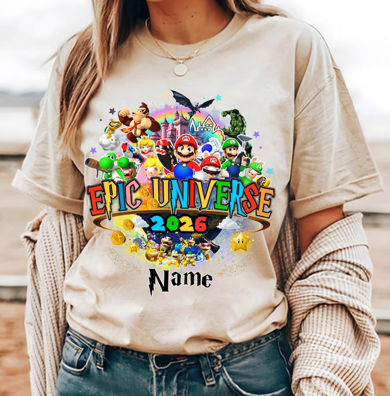 Pu&ograve; includere: T-shirt beige con una grafica colorata di personaggi di videogiochi, un arcobaleno e la scritta "Epic Universe 2026". Il design include uno spazio per un nome personalizzato. La maglietta ha maniche corte e girocollo.