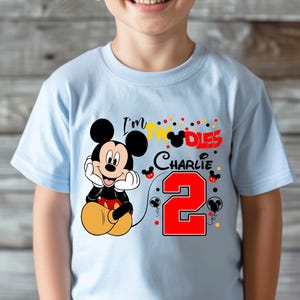May include: Light blue t-shirt featuring a smiling Mickey Mouse graphic. The shirt has the text "I'm 2" with the name "Charlie" and a large red number 2. The design includes colorful polka dots and Mickey Mouse silhouettes.