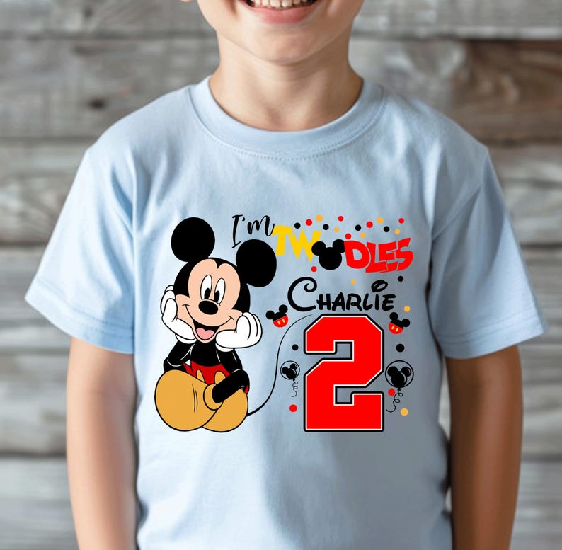 May include: Light blue t-shirt featuring a smiling Mickey Mouse graphic. The shirt has the text "I'm 2" with the name "Charlie" and a large red number 2. The design includes colorful polka dots and Mickey Mouse silhouettes.