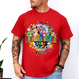 Pu&ograve; includere: T-shirt rossa con una grafica colorata. Il design include il testo "EPIC UNIVERSE 2026" e vari personaggi dei cartoni animati. La parola "Name" &egrave; sotto il testo principale, con stelle e un castello.