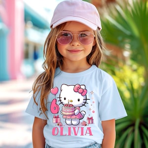 May include: A light blue t-shirt with a Hello Kitty design holding a birthday cake with the number 6. The name "OLIVIA" is printed below the graphic in pink. The shirt is worn by a young girl wearing a pink cap and sunglasses.