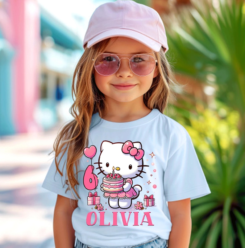 May include: A light blue t-shirt with a Hello Kitty design holding a birthday cake with the number 6. The name "OLIVIA" is printed below the graphic in pink. The shirt is worn by a young girl wearing a pink cap and sunglasses.