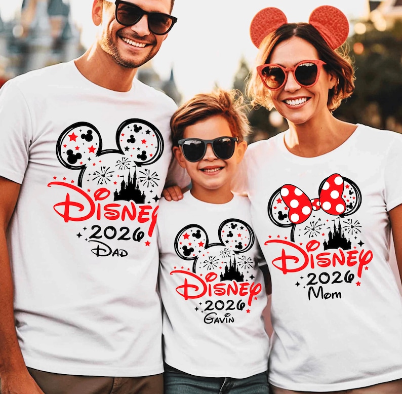 May include: Three white t-shirts featuring a black Mickey Mouse ear silhouette filled with red stars and fireworks. The design includes the red text Disney, the year 2026, and the names Dad, Gavin, and Mom. The shirts feature a castle graphic.