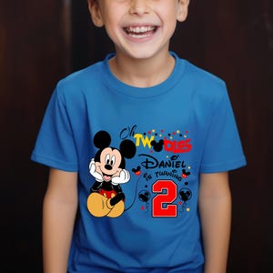 May include: A blue t-shirt featuring Mickey Mouse and the text "Oh TWO-DIES Daniel is turning 2". Mickey Mouse is in a seated position with his hands under his chin. The number 2 is in red with black balloons.