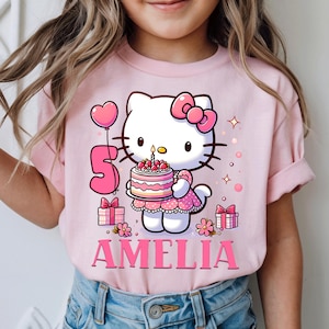 May include: A pink t-shirt with a Hello Kitty graphic holding a birthday cake with a lit candle. The number 5 and the name "Amelia" are displayed. Pink gifts and a heart-shaped balloon complete the design.
