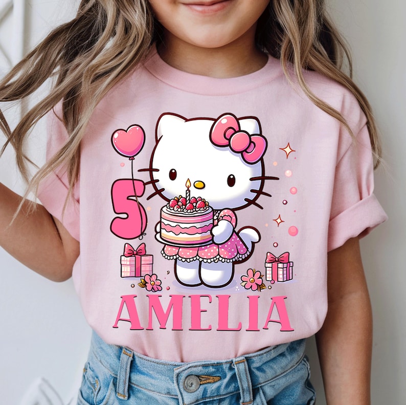 May include: A pink t-shirt with a Hello Kitty graphic holding a birthday cake with a lit candle. The number 5 and the name "Amelia" are displayed. Pink gifts and a heart-shaped balloon complete the design.