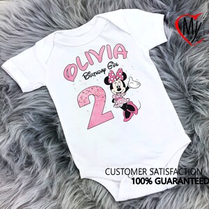 Puede incluir: Body de beb&eacute; blanco con un gr&aacute;fico de Minnie Mouse rosa. El body presenta el nombre "OLIVIA" y el texto "Birthday Girl" en rosa, junto con el n&uacute;mero "2". El body tambi&eacute;n tiene el texto "SATISFACCI&Oacute;N DEL CLIENTE 100% GARANTIZADA".