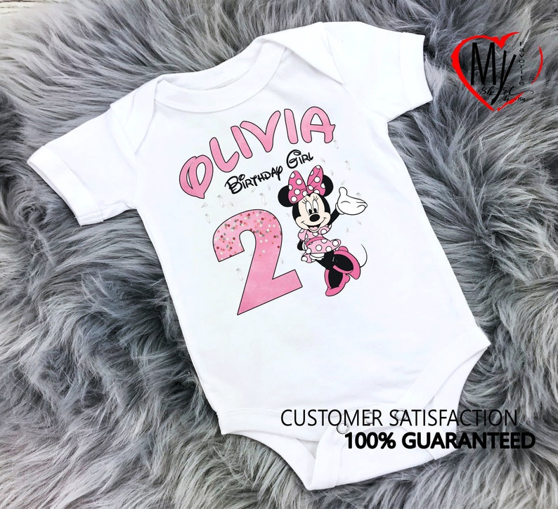 Puede incluir: Body de beb&eacute; blanco con un gr&aacute;fico de Minnie Mouse rosa. El body presenta el nombre "OLIVIA" y el texto "Birthday Girl" en rosa, junto con el n&uacute;mero "2". El body tambi&eacute;n tiene el texto "SATISFACCI&Oacute;N DEL CLIENTE 100% GARANTIZADA".