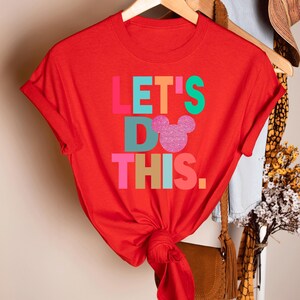 May include: A red t-shirt with the phrase "LET'S DO THIS." in colorful block letters. A glittery pink Mickey Mouse head replaces the "O" in "DO." The shirt is hanging on a wooden hanger.