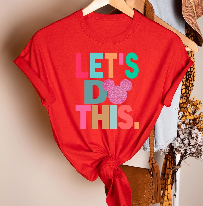 May include: A red t-shirt with the phrase "LET'S DO THIS." in colorful block letters. A glittery pink Mickey Mouse head replaces the "O" in "DO." The shirt is hanging on a wooden hanger.